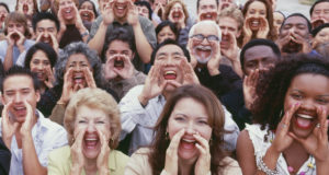 Panoramic shot of crowd shouting with hands on their faces.