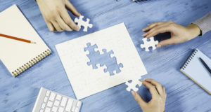 An image of a jigsaw puzzle with missing pieces which are being held by three hands surrounding the puzzle.