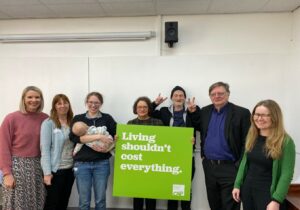 Picture of seven adults smiling and one holding a baby with an adult holding a sign. Sign says "Living shouldn't cost everything"